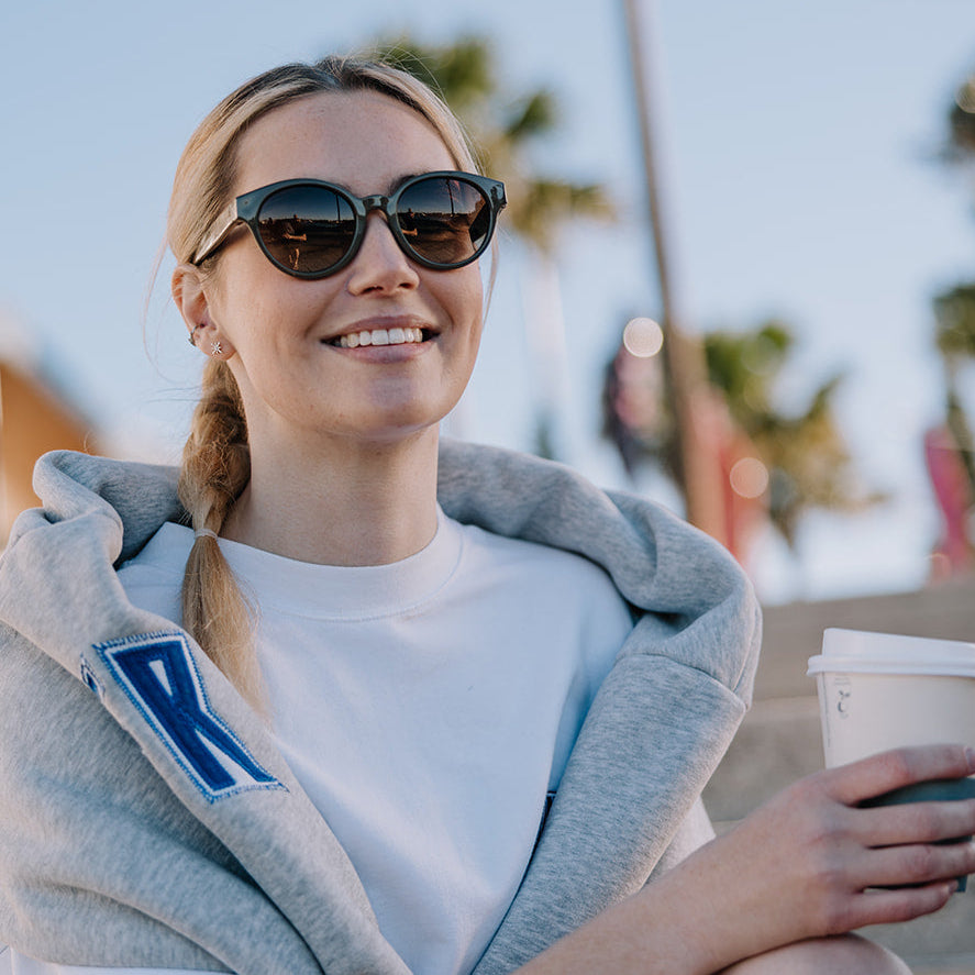 A woman sitting outdoors on stone steps, wearing a white T-shirt, a light gray hoodie draped over her shoulders, and ugly fish women's sunglasses, holding a takeaway coffee cup. Palm trees and a blue sky are blurred in the background.
