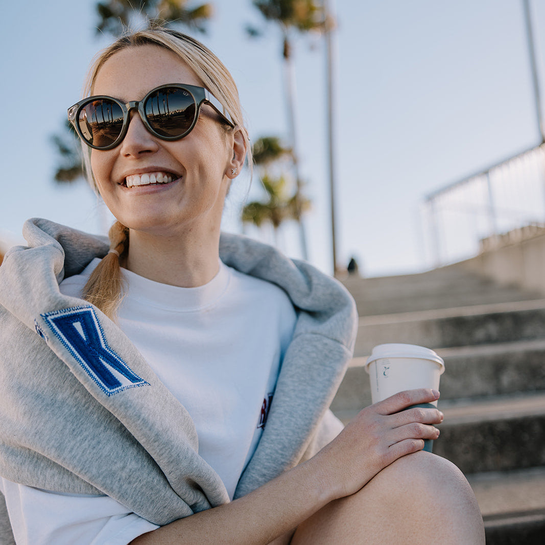 A woman sitting outdoors on concrete steps wearing ugly fish women's sunglasses, a white T-shirt, and a light gray hoodie draped over her shoulders. She holds a white paper coffee cup, with palm trees and a railing blurred in the background.