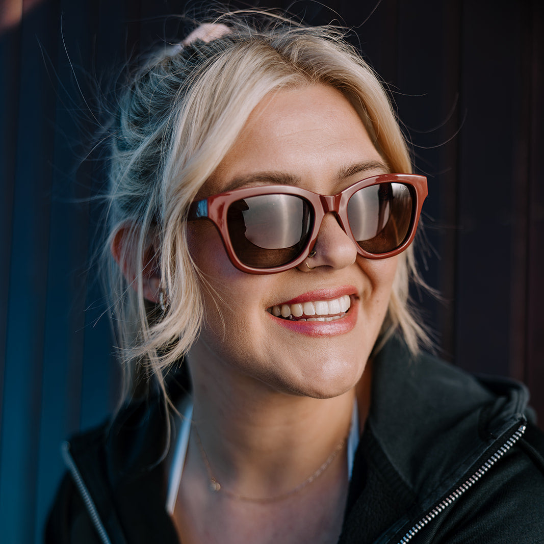A smiling blonde woman wearing Ugly Fish Women's sunglasses with reddish-brown frames and dark lenses, paired with a black zip-up hoodie against a dark background.