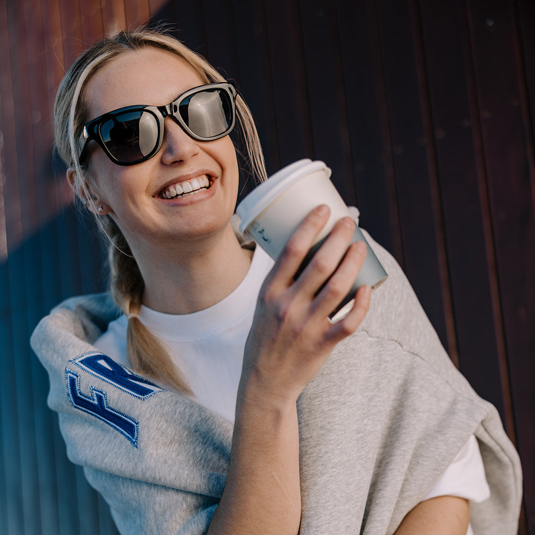 A smiling woman wearing black Ugly Fish women's sunglasses, a white t-shirt, and a gray knit sweater draped over her shoulders, holding a takeaway coffee cup against a dark wooden background.
