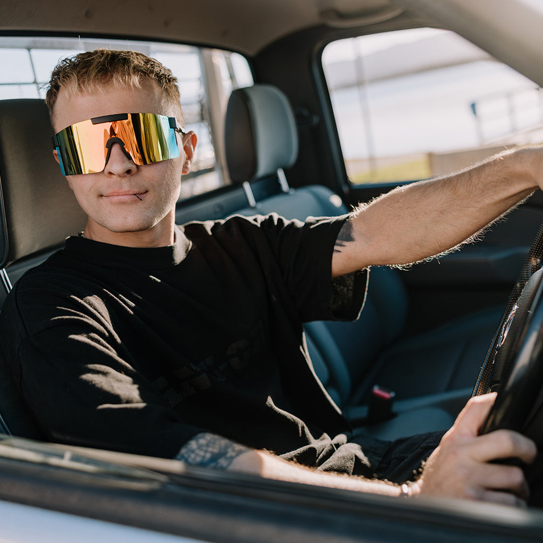 A man wearing ugly fish reflective shield sunglasses and a black t-shirt sits in the driver's seat of a car, holding the steering wheel.