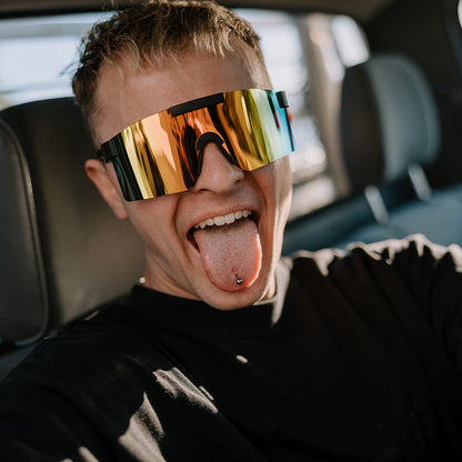 A man wearing ugly fish shield sunglasses with a reflective gold-to-blue mirrored lens, sticking his tongue out while sitting in a car.