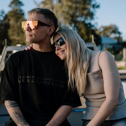 A couple sitting together outdoors in the sun, with the man wearing a black graphic t-shirt and black pants, and the woman wearing a beige tank top and jeans.