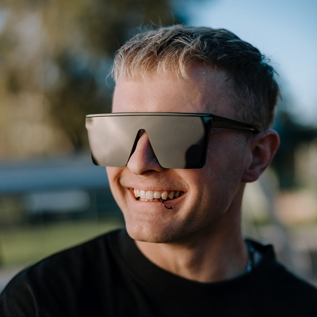 A smiling man wearing ugly fish shield-style sunglasses with a dark frame and reflective lenses, outdoors in warm natural light.