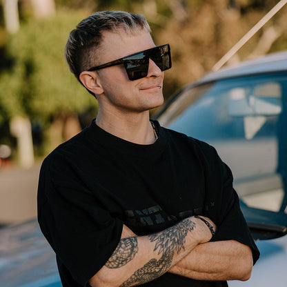 A man with short hair and tattoos wearing black ugly fish sunglasses and a black t-shirt leans against a white car outdoors.