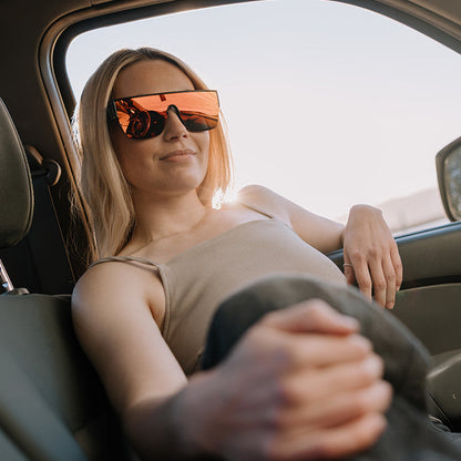 A woman wearing orange-tinted ugly fish sunglasses and a beige tank top reclines on the back seat of a car interior.