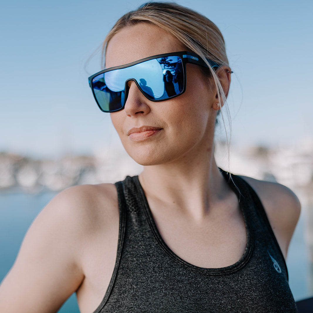 A woman outdoors wearing ugly fish mirrored blue sunglasses and a dark gray athletic tank top, with a blurred waterfront background.