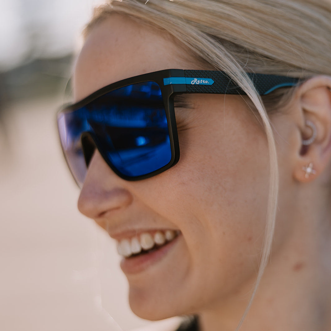 A close-up side profile of a smiling blonde woman wearing ugly fish blue mirrored sunglasses with a black frame and a small blue brand logo on the temple.