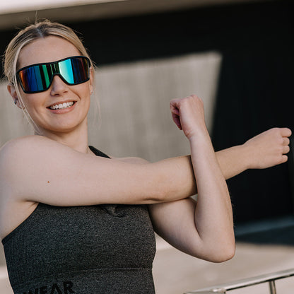A woman in a dark gray sports bra and mirrored ugly fish sunglasses stretches her arm, standing outdoors in front of a modern concrete wall.