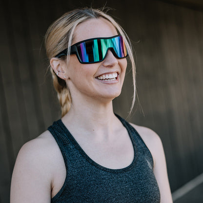A woman wearing oversized shield-style ugly fish sunglasses with reflective rainbow lenses and a dark athletic tank top, standing against a dark wooden wall.