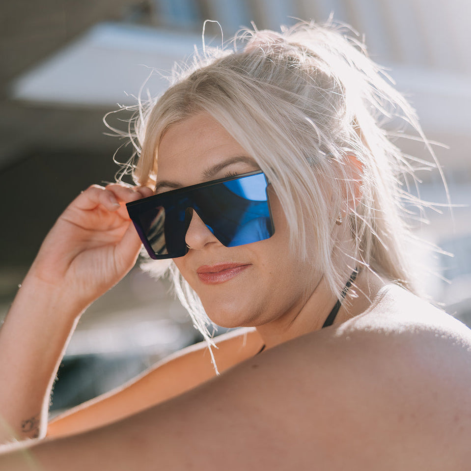 A woman with blonde hair wearing ugly fish black sunglasses with reflective blue lenses, holding them up to her eyes in bright outdoor light.