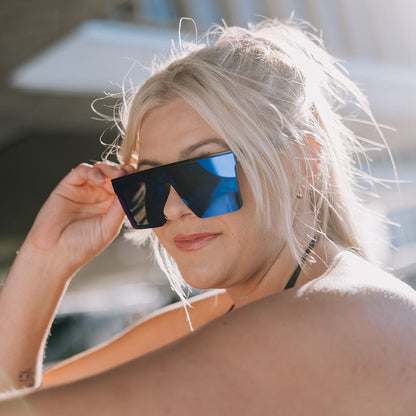 A woman with blonde hair wearing ugly fish black sunglasses with reflective blue lenses, holding them up to her eyes in bright outdoor light.