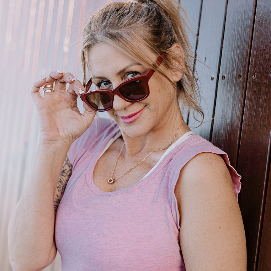 A woman wearing a pink sleeveless top and Ugly Fish women's sunglasses leans against a dark brown wooden wall.