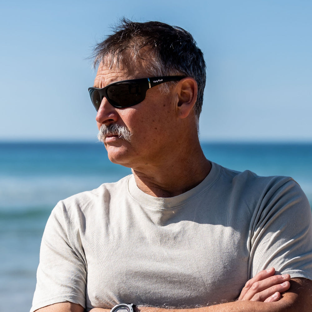 Man standing on a beach with arms crossed, wearing Ugly Fish Polarised sunglasses and a watch.