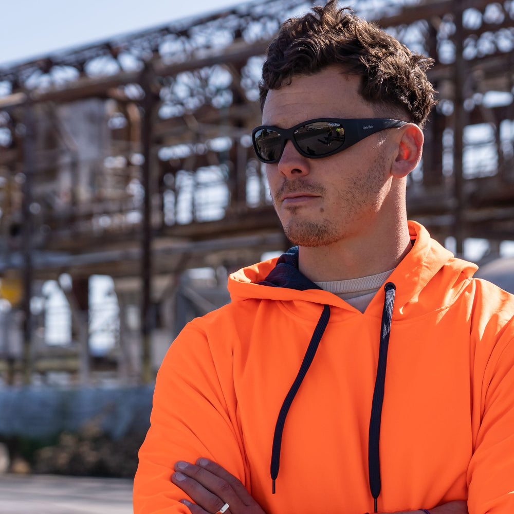 Person wearing an orange hoodie and Ugly Fish Safety sunglasses with a industrial background