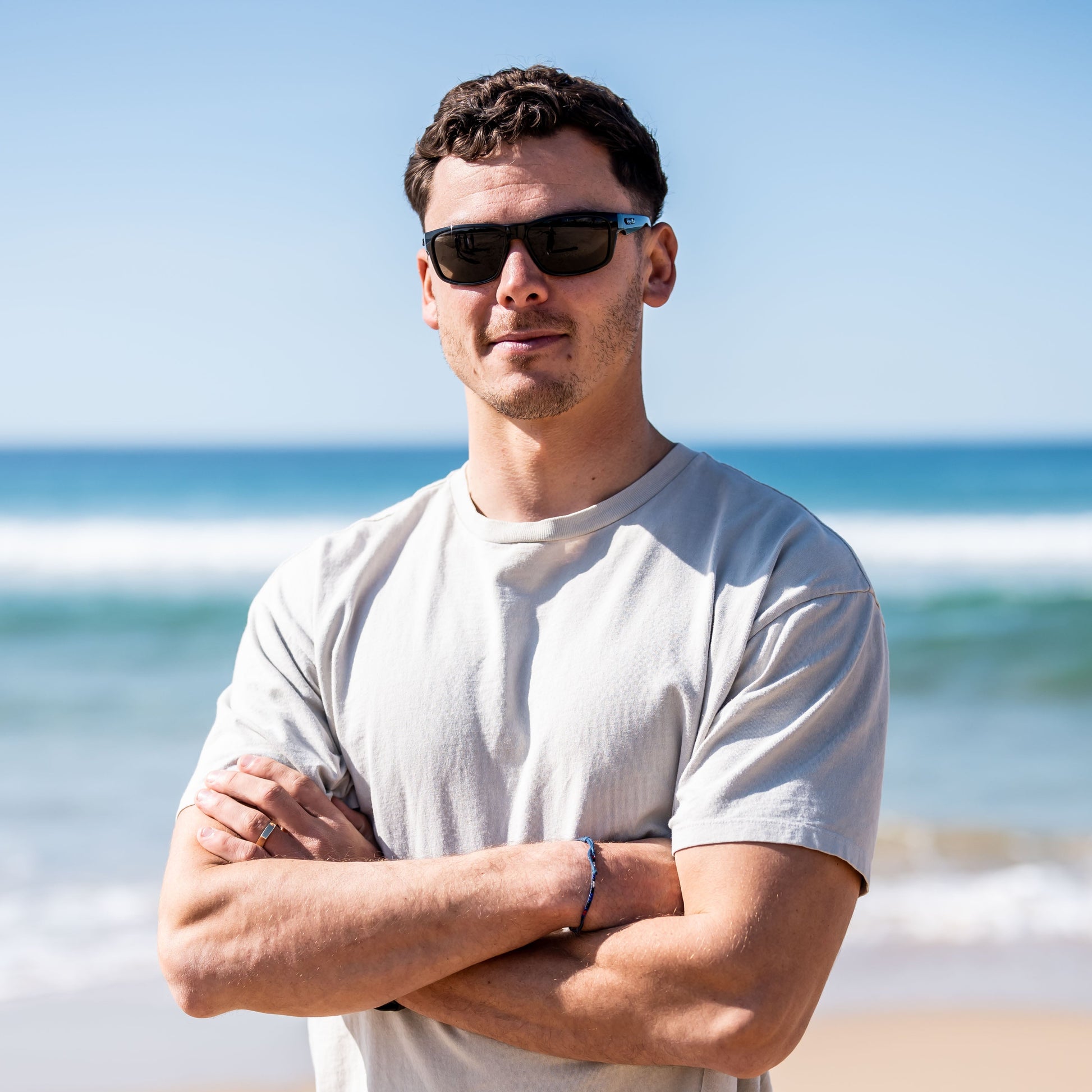 Man wearing Ugly Fish Xenon Bifocal sunglasses and a white t-shirt standing on a beach with ocean waves in the background