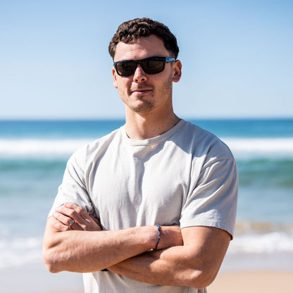 Man wearing Ugly Fish Xenon Bifocal sunglasses and a white t-shirt standing on a beach with ocean waves in the background