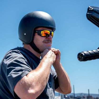 Man wearing a helmet and Ugly Fish motorcycle sunglasses by a waterfront with clear blue sky