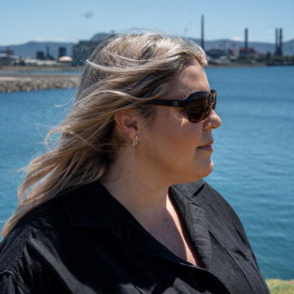 Woman wearing Ugly Fish polarised sunglasses and a black shirt by a body of water with industrial structures in the background.