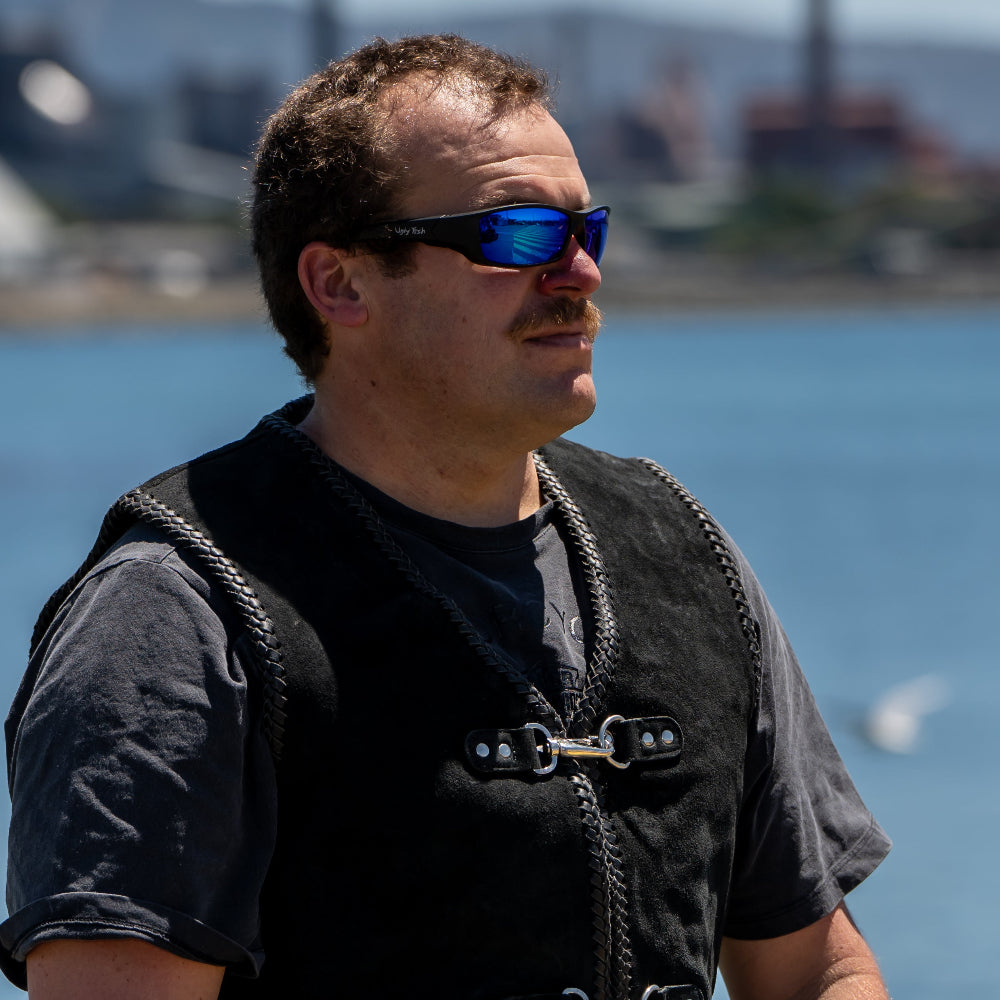 Man wearing Ugly Fish motorcycle sunglasses and a black vest standing by a body of water with mountains in the background.