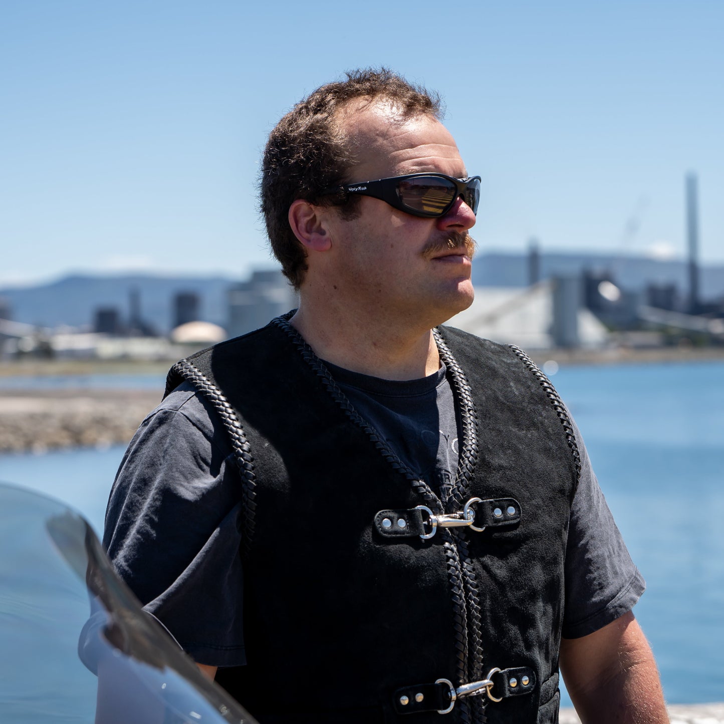Man wearing Ugly Fish Ultimate Motorcycle sunglasses and a black vest standing by a waterfront with industrial buildings in the background.