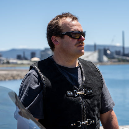 Man wearing Ugly Fish Ultimate Motorcycle sunglasses and a black vest standing by a waterfront with industrial buildings in the background.