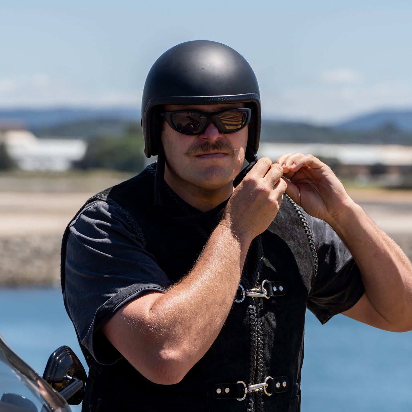 Man wearing a black helmet and Ugly Fish Ultimate Motorcycle sunglasses standing next to a motorcycle by a waterfront.