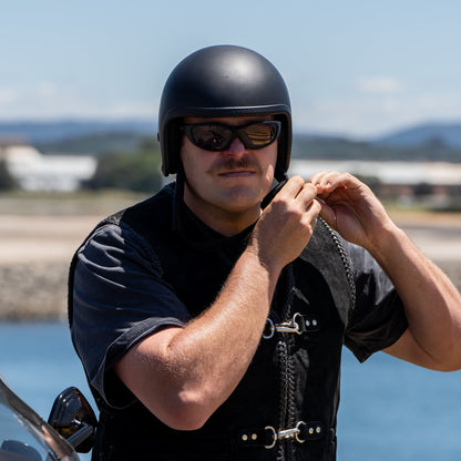 Man wearing a black helmet and Ugly Fish Ultimate Motorcycle sunglasses standing next to a motorcycle by a waterfront.