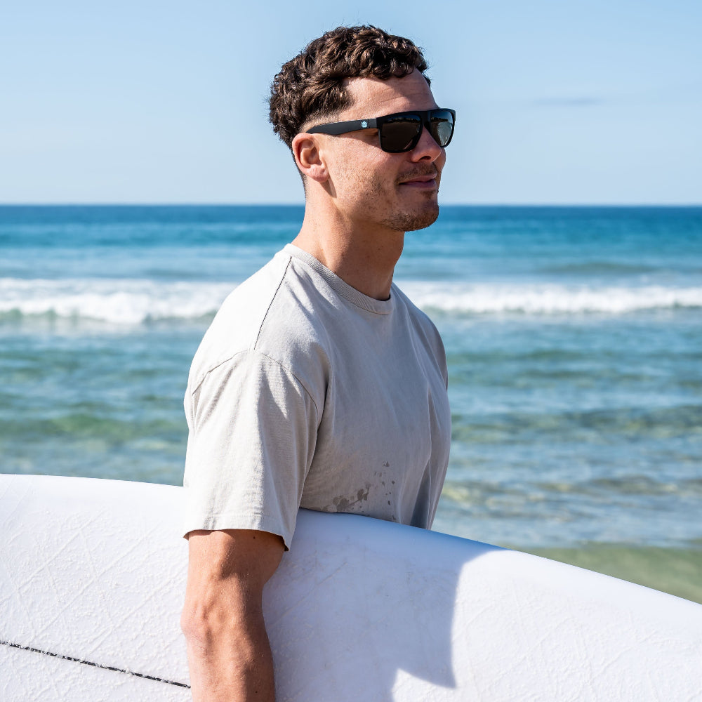 Man holding a surfboard wearing Ugly Fish floating sunglasses with ocean and clear sky in the background