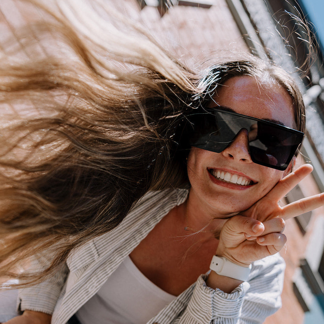 Woman making a peace sign with Rogue sunglasses on, standing next to a wall.